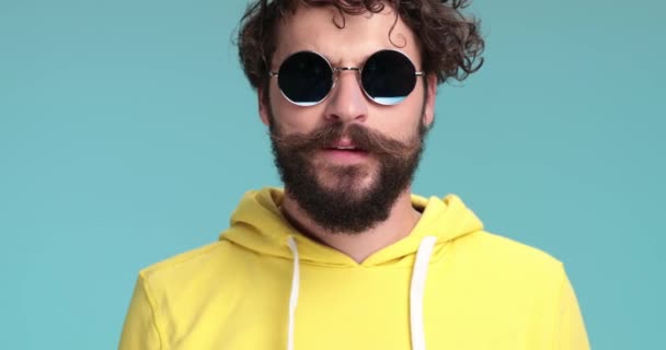 cool handsome curly hair man wearing yellow sweatshirt taking glasses off, approaching and observing, covering mouth with hand and being shocked in studio - Footage, Video
