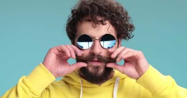 curly hair man in yellow sweatshirt arranging moustache, wearing sunglasses and making thumbs up gesture while confidently smiling on blue background - Footage, Video