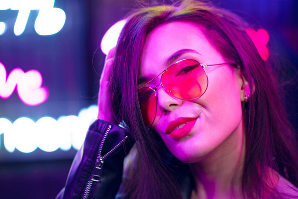 Fashion portrait of a young woman in sunglasses posing near neon signs in night club - Фото, зображення