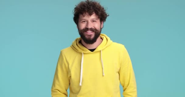 chladný ležérní hipster s dlouhým vousem a knírkem, oblečený ve žluté mikině, ukazující prsty a usmívající se na modrém pozadí ve studiu - Záběry, video