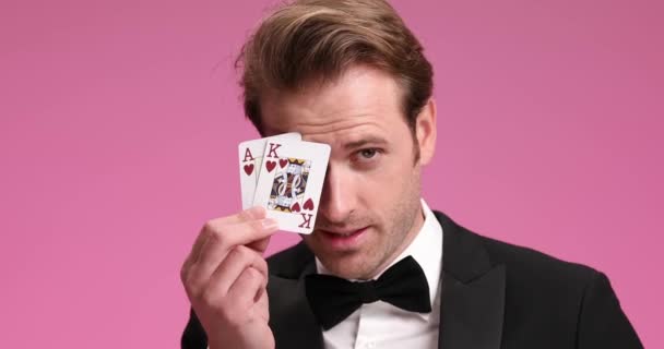zelfverzekerde jonge gokker dekking oog met aas en koning, opkijken en glimlachen terwijl het tonen van kaarten tegen roze achtergrond in de studio - Video