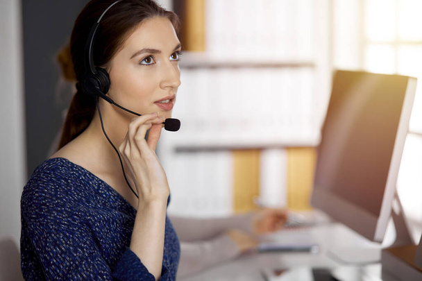 Asian or Latin american businesswoman talking by headset in sunny office. Call center and diverse people group in business - Fotoğraf, Görsel