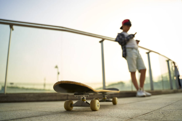 十代のアジアのスケートボーダー少年は休みながら携帯電話を見て、スケートボードに焦点を当て - 写真・画像