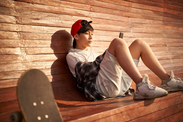adolescente asiático skateboarder chico buscando en celular mientras descansando - Foto, imagen