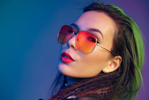 Asian girl in sunglasses and black leather jacket posing against dark blue background - Фото, изображение