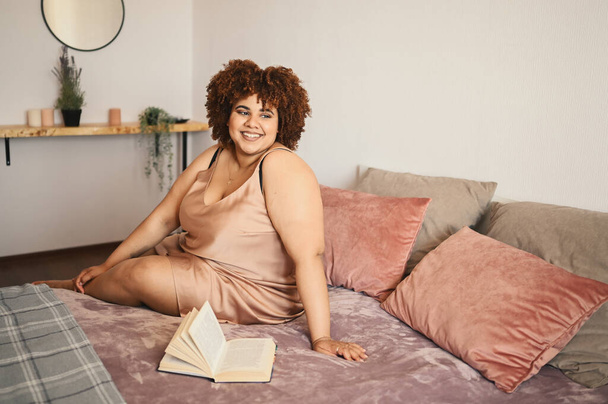 Gyönyörű görbe plusz méret Afrikai fekete nő afro haj fekszik az ágyon selyem por rózsaszín ruha hangulatos hálószoba belsőépítészeti. A test tökéletlensége, a test elfogadása, a test pozitív és sokszínű koncepciója - Fotó, kép