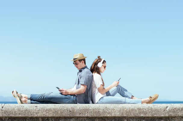 Couple assis dos à dos tout en vérifiant leurs smartphones sur la côte par une journée ensoleillée. Espace de copie bleu au ciel. - Photo, image