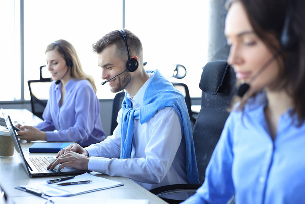 Smiling male call-center operator with headphones sitting at modern office with collegues on the backgroung, consulting online - Foto, Imagem