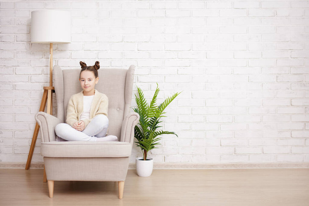 linda chica preadolescente sentada en sillón sobre fondo de pared de ladrillo blanco con espacio de copia - Foto, imagen