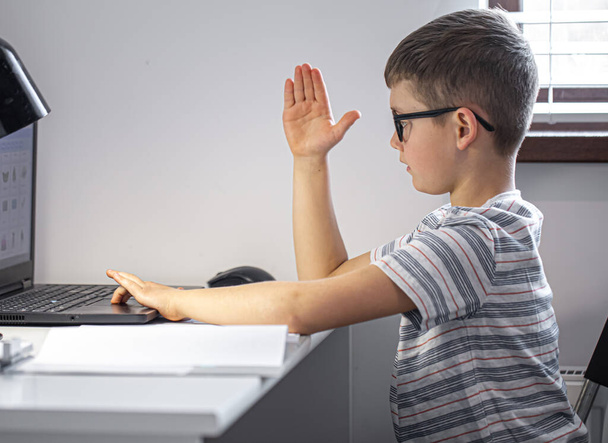 Um estudante do ensino fundamental com óculos senta-se em uma mesa com um laptop, aprende remotamente, levanta a mão em uma lição on-line. - Foto, Imagem