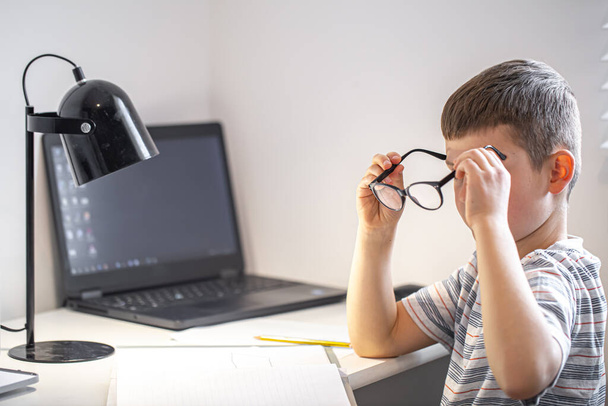 Egy általános iskolás diák ül egy asztalnál egy laptoppal, szemüveget tartva a kezében.. - Fotó, kép