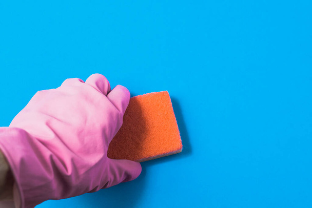 A hand in a pink rubber glove holds a orange sponge. Blue background. - Photo, Image