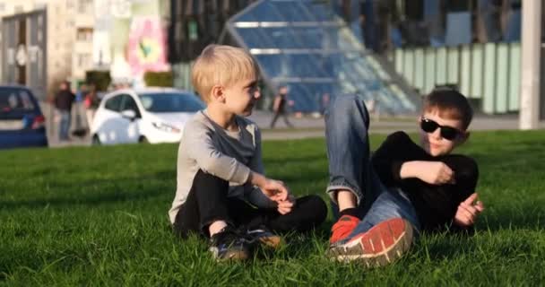 Jongen ligt op het gazon en praat met zijn jongere broer. Vrienden communiceren buiten. Zomertijd, vakantiedagen - Video