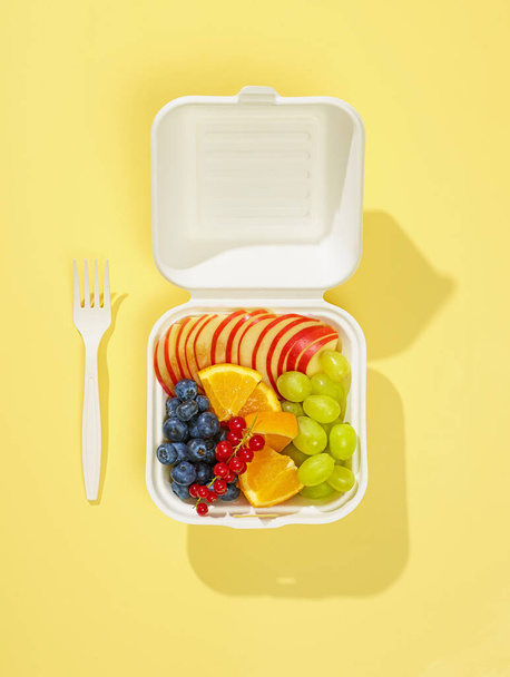 fruta fresca en rodajas y bayas en caja blanca para llevar aislada sobre fondo amarillo, vista superior - Foto, Imagen