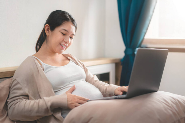 Mutlu Asyalı hamile kadın gülümsüyor ve bilgisayarını yatak odasında çalışmak için kullanıyor. Uzaktan kumanda, online alışveriş, kadın hamilelik konsepti, yeni normal ve ev konseptinden çalışma.. - Fotoğraf, Görsel
