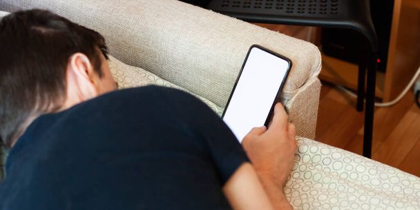 A man in a blue T-shirt lies on the couch and looks at his smartphone. The concept of laziness and the use of gadgets while lying on the couch. White Ecoran Gadget - Photo, Image