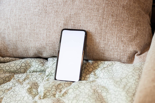 ソファの上のクッションの上に白い画面を持つフレームのないスマートフォン。枕に電子機器を使うという考え方 - 写真・画像