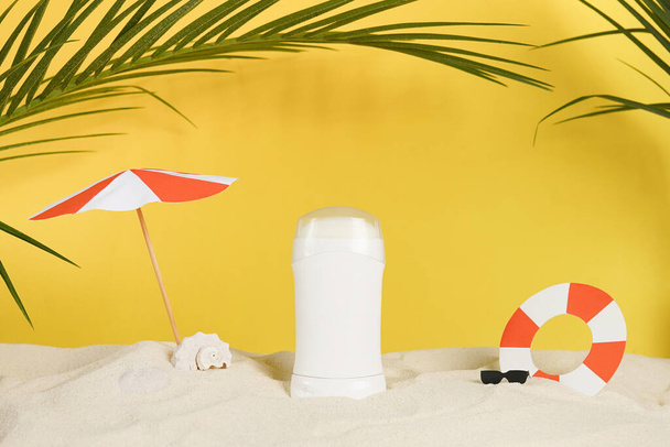 déodorant blanc ou antitranspirant sur fond ensoleillé d "été avec sable sur la plage au bord de la mer, feuilles de palmier vertes et accessoires en papier - Photo, image
