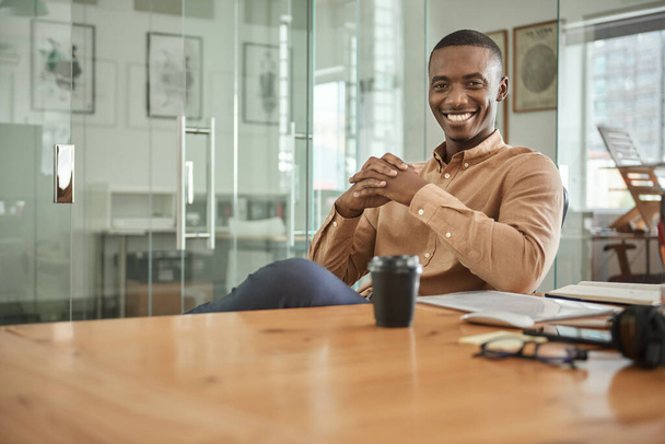 Portrait of a smiling young African businessman sitting at his desk in a modern office - Fotografie, Obrázek