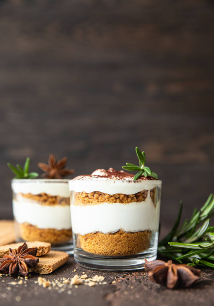 Layered dessert in glass jar with cookie crumble and whipped cream decorated with rosemary and anise, dark background. No bake cheesecake, trifle or pudding. - Фото, зображення