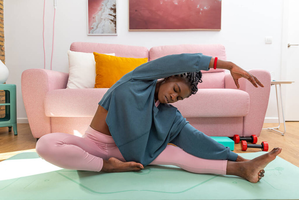 Черная женщина сидит на коврике и растягивает ноги, делая боковой изгиб во время тренировки дома - Фото, изображение