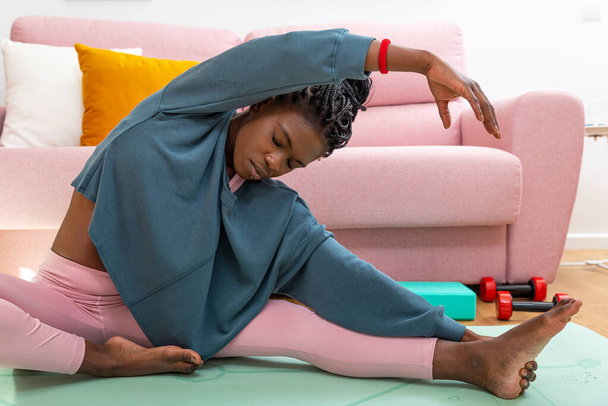 Black woman sitting on mat and stretching legs while doing side bend during training at home - Photo, Image