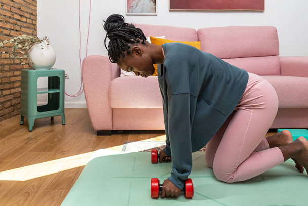 Side view of concentrated black sportswoman exercising with dumbbells on mat during workout at home - Foto, afbeelding