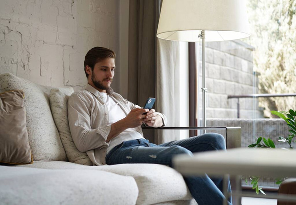 Handsome man in basic t-shirt smiling and holding mobile phone in hands while sitting on couch in living room. - Фото, изображение