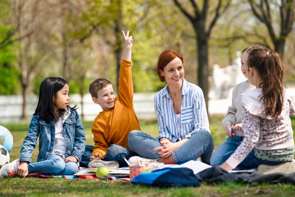 Teacher with small children sitting outdoors in city park, learning group education concept. - 写真・画像