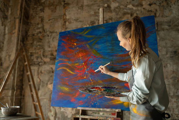 Female artist in front of an easel, painting with a brush, using oil or acrylic paint - Foto, Imagen