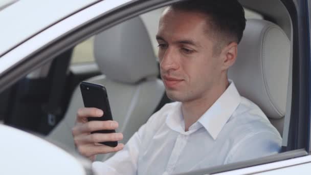 Close-up portrait of smiling male businessman sitting at the wheel of a car and using smartphone. - Footage, Video