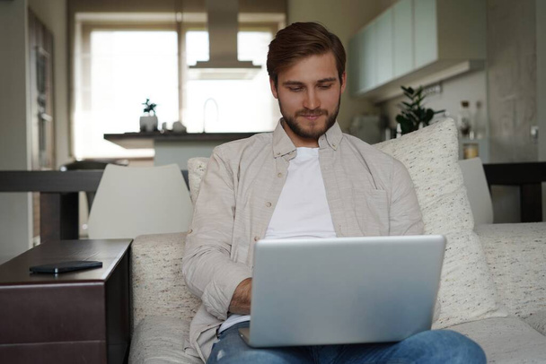 Man using laptop, sitting on couch at home, chatting or shopping in internet, freelancer working online - Photo, image