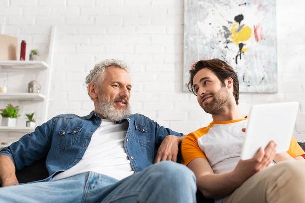 Man holding blurred digital tablet near mature father on couch  - Photo, Image
