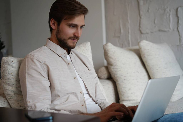 Man using laptop, sitting on couch at home, chatting or shopping in internet, freelancer working online - Photo, Image