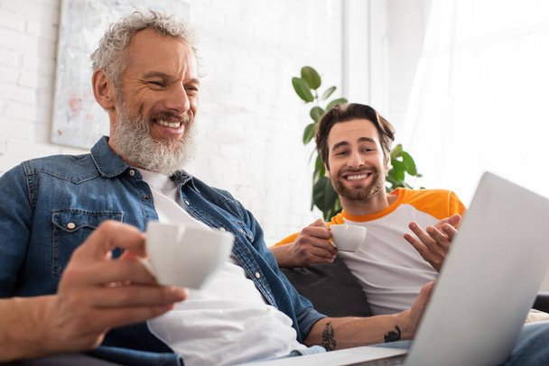 Smiling man holding coffee near son and blurred laptop  - Foto, afbeelding