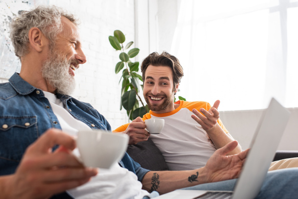 Smiling man with coffee pointing at blurred laptop near father at home  - Photo, Image