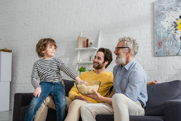 Мужчины с попкорном сидят на диване рядом с ребенком  - Фото, изображение