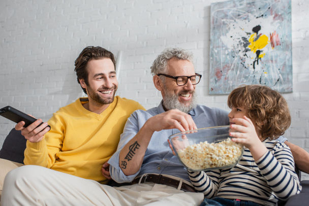Smiling man with remote controller sitting near father and son with popcorn on couch  - Photo, Image