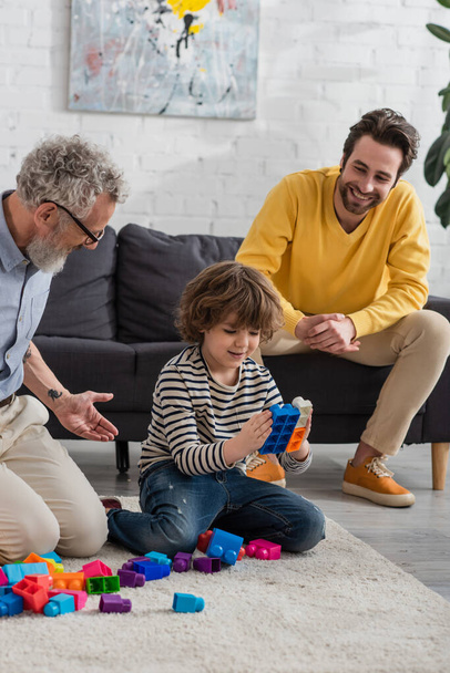 Smiling kid playing building blocks near father and grandfather at home  - Photo, Image