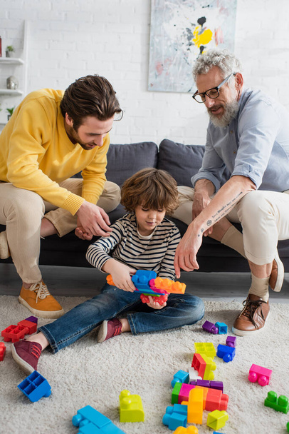 Volwassen man en zoon zitten in de buurt van kleinzoon met bouwstenen op de vloer  - Foto, afbeelding