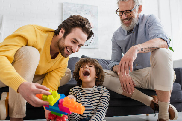 Возбужденный мальчик сидит рядом с улыбающимися родителями с размытыми кирпичами дома  - Фото, изображение