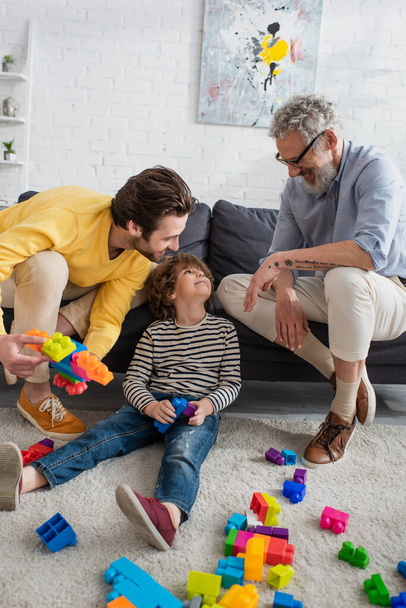 Positive parents looking at kid near building blocks on carpet  - Photo, Image