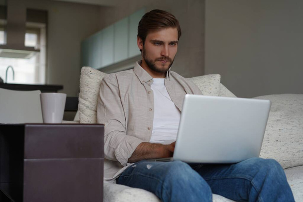 Man using laptop, sitting on couch at home, chatting or shopping in internet, freelancer working online - Foto, Imagem