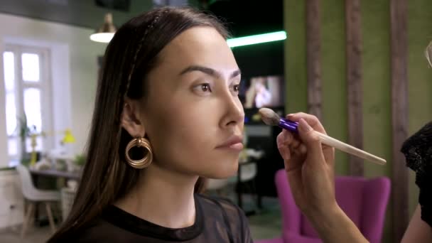 makeup artist applies base tone cream on girls face using brush in beauty salon - Footage, Video