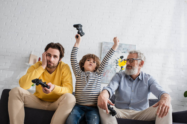 KYIV, UKRAINE - APRIL 12, 2021: Low angle view of excited kid with joystick near sad parents on couch  - Photo, Image