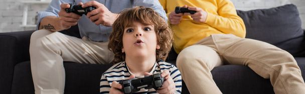KYIV, UKRAINE - KWIECIEŃ 12, 2021: Podekscytowany chłopak grający w grę wideo z rozmytymi rodzicami, baner  - Zdjęcie, obraz