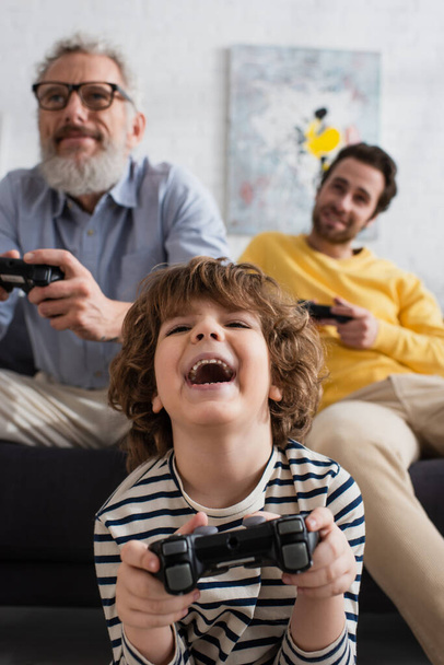 KYIV, UKRAINE - APRIL 12, 2021: Excited kid playing video game near blurred parents  - Photo, Image