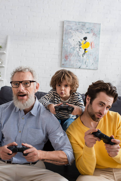 KYIV, UKRAINE - APRIL 12, 2021: Parent and granddad playing video game with kid at home  - Photo, image