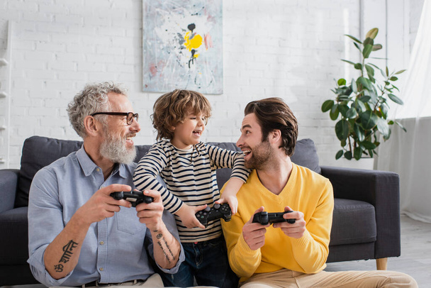 KYIV, UKRAINE - APRIL 12, 2021: Cheerful kid looking at father with joystick near grandfather  - Foto, imagen