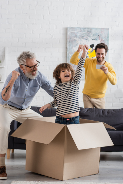 Excited kid showing yes gesture in carton box near parent with smartphone and grandfather  - Fotografie, Obrázek